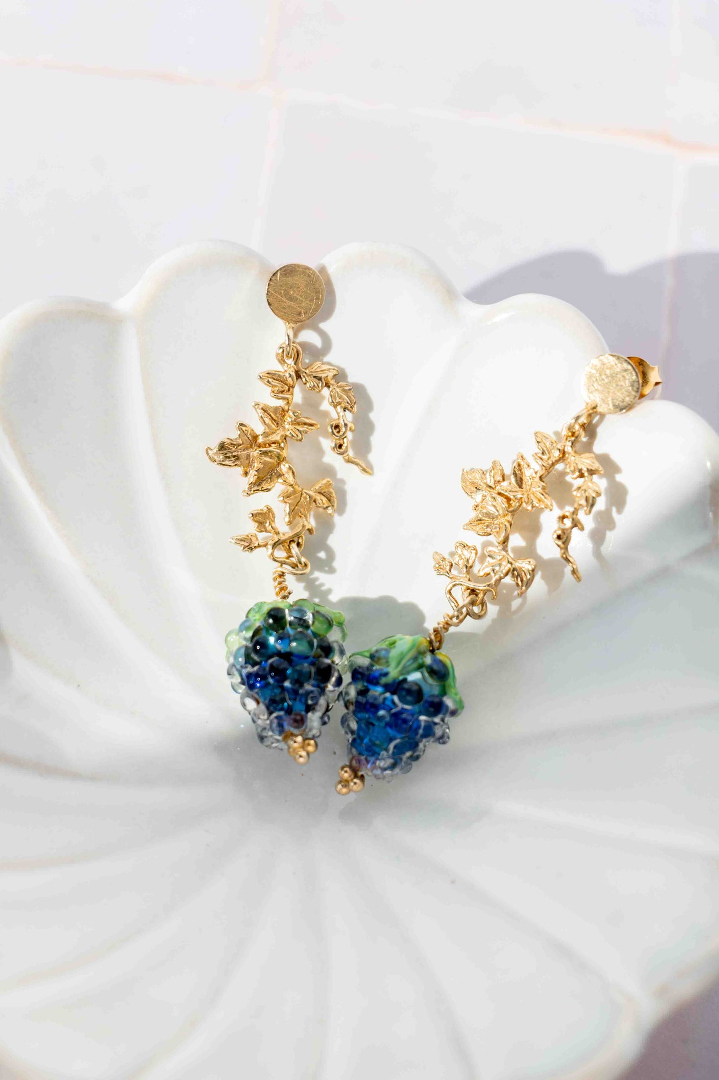 Gold pair of earrings with blue and green grape fruits on a white dish