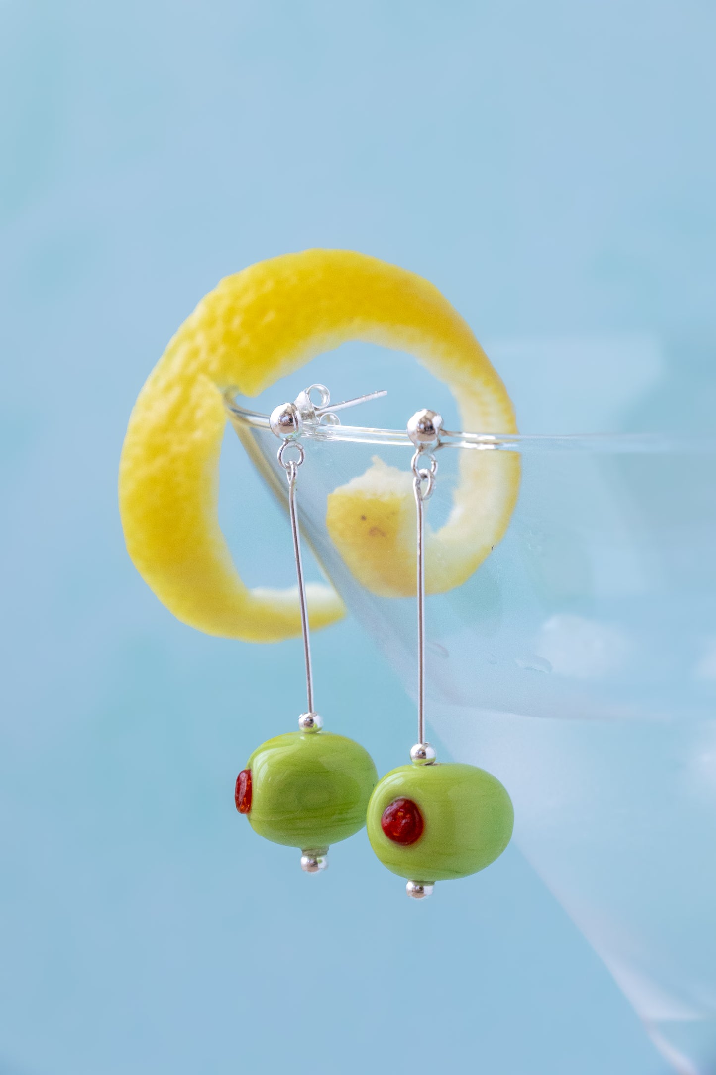 Green olive dangle earrings with small red eye details on silver hooks, displayed against a light blue background with a yellow lemon twist garnish.