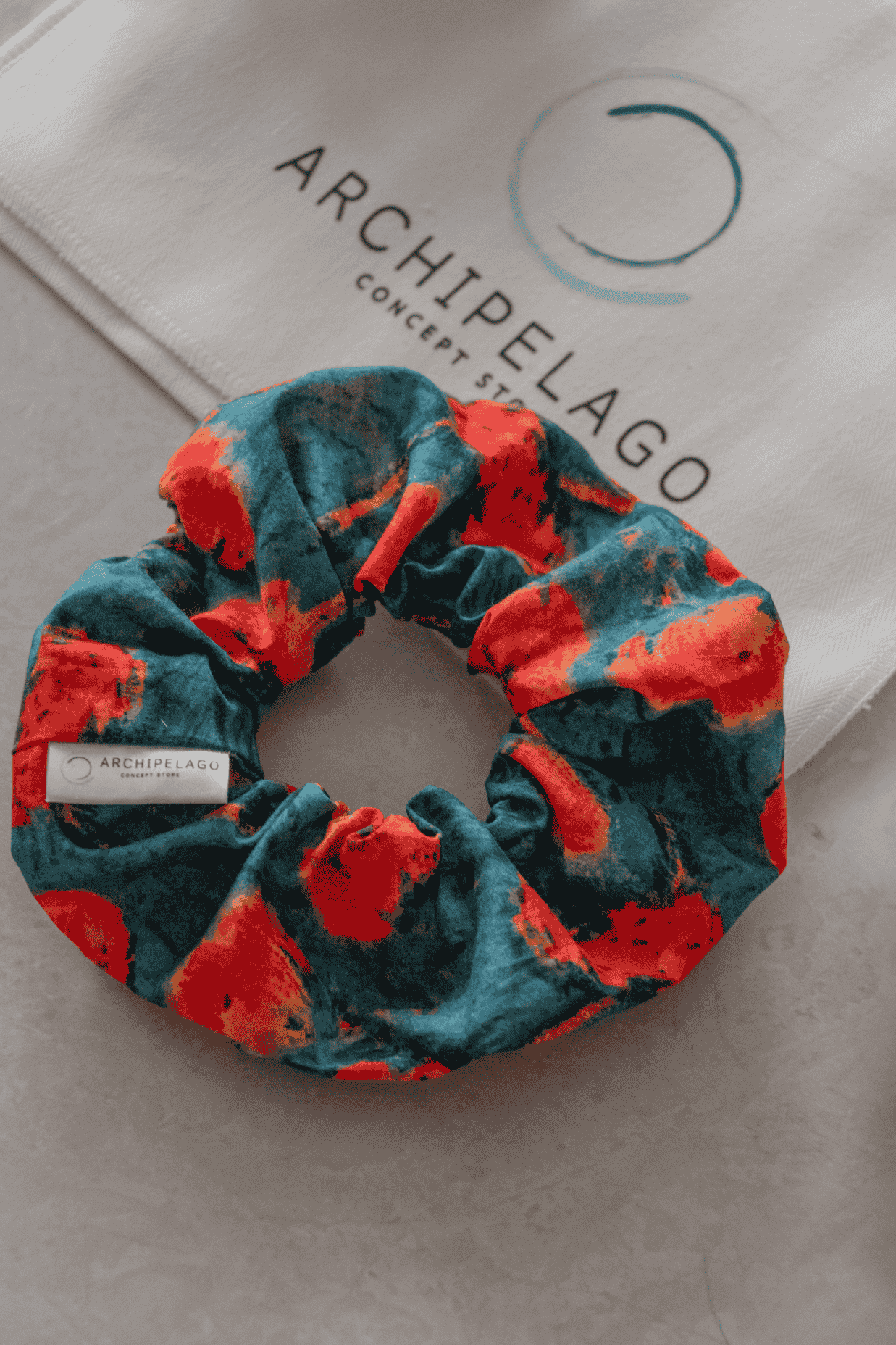 Scrunchie with red and blue pattern on a white surface with 'Archipelago' branding.