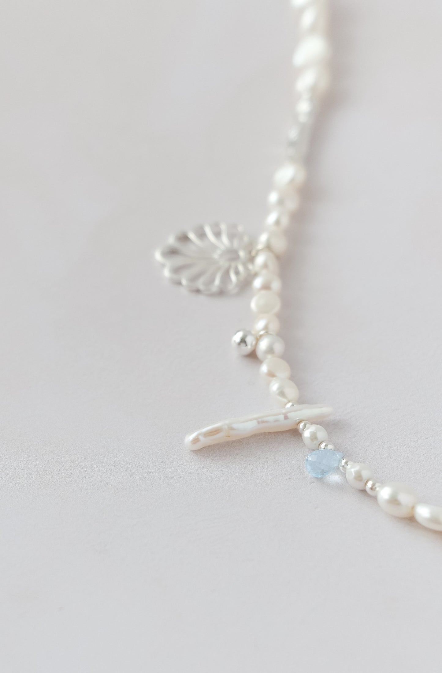 A delicate silver necklace with white pearl-like beads, a silver filigree heart charm, a small silver cross charm, and a tiny light blue faceted bead, shown on a light gray background.