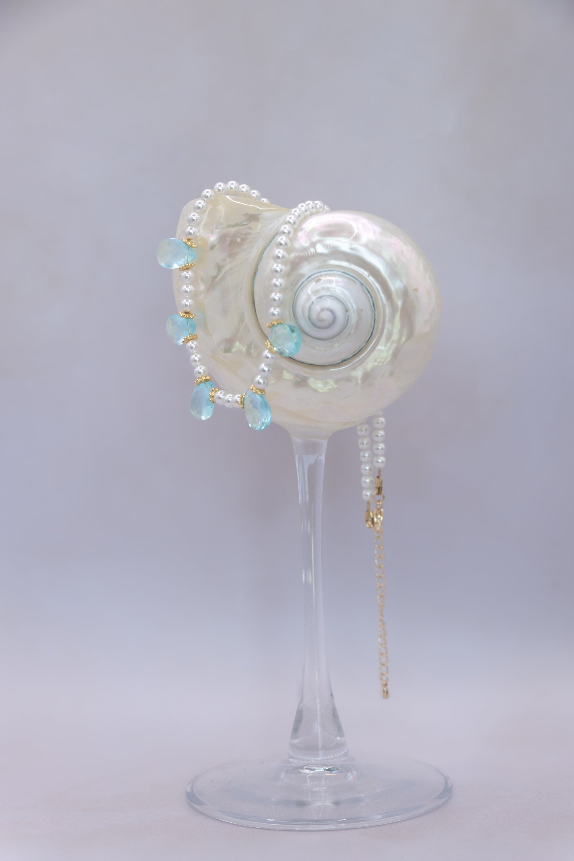 A pearl and light blue bead necklace draped over a white seashell-shaped display stand on a clear glass pedestal, photographed against a soft light gray studio background.