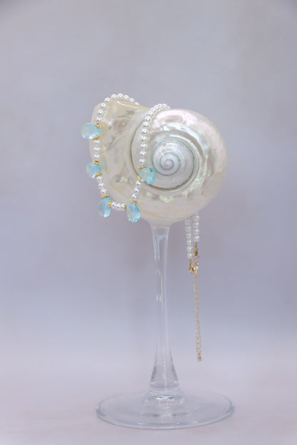 A pearl and light blue bead necklace draped over a white seashell-shaped display stand on a clear glass pedestal, photographed against a soft light gray studio background.