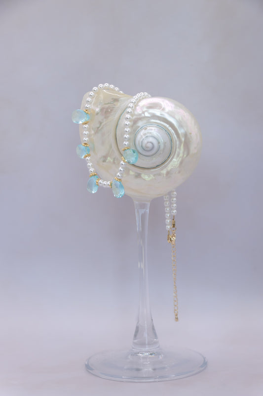 A pearl and light blue bead necklace draped over a white seashell-shaped display stand on a clear glass pedestal, photographed against a soft light gray studio background.