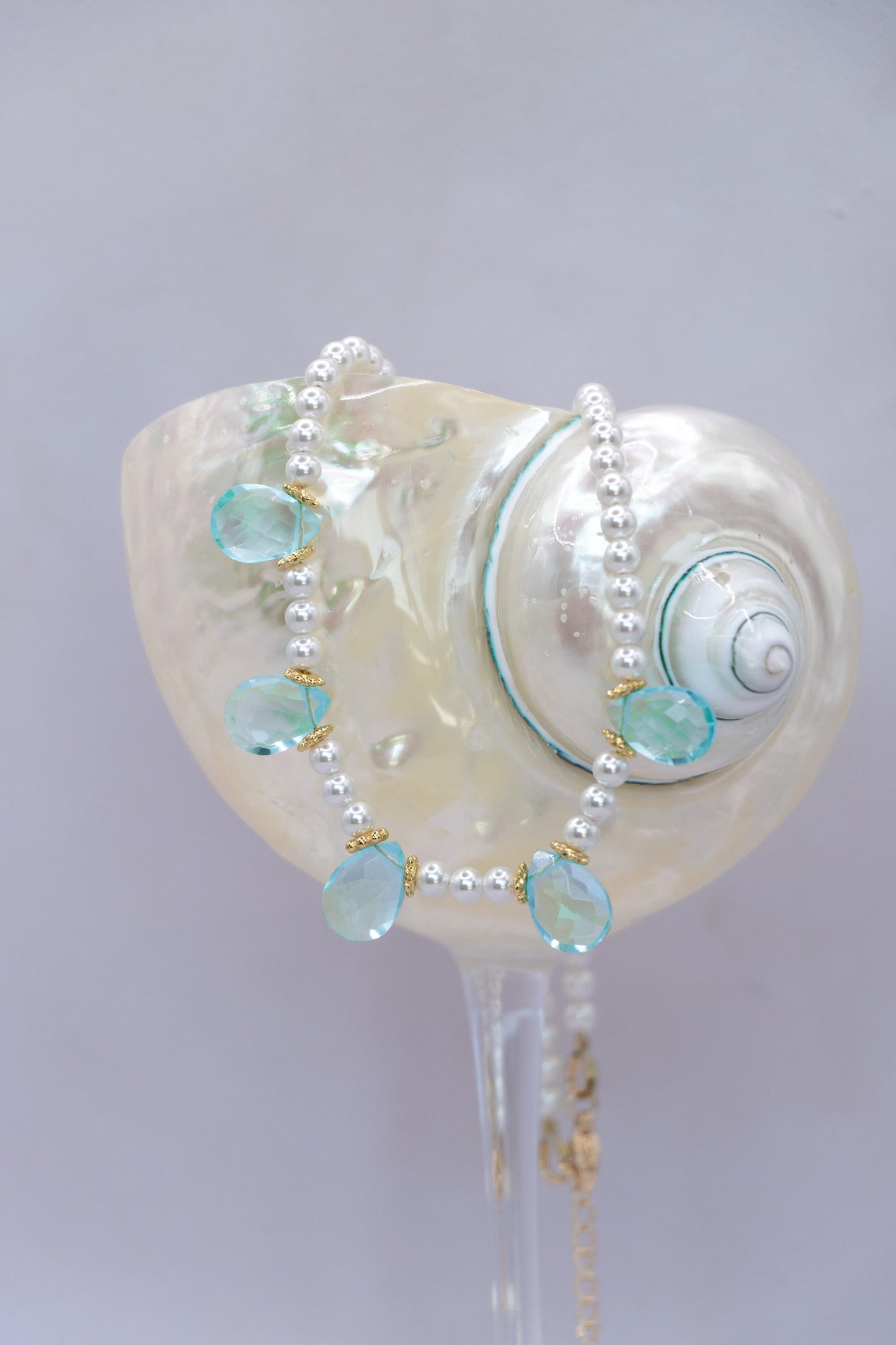 A pearl beaded necklace with light blue teardrop gemstones and gold accents, draped over a large white seashell display stand.