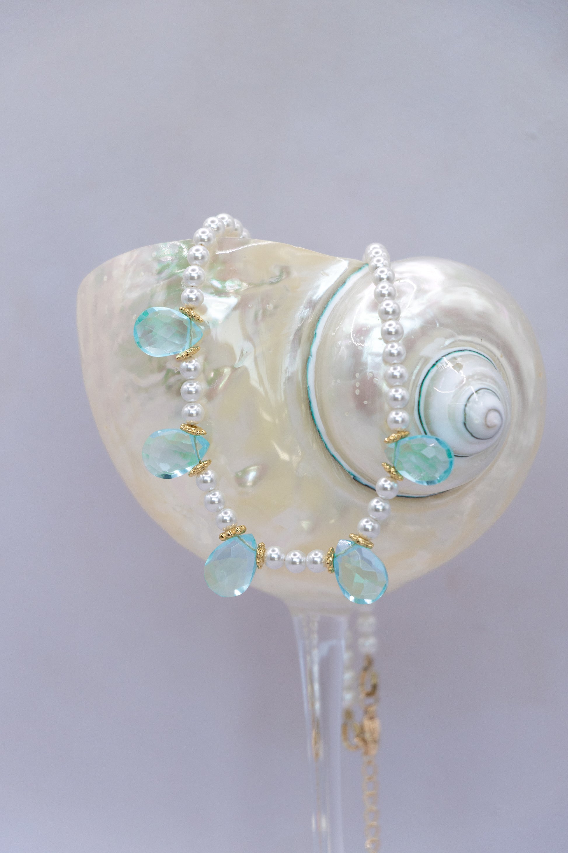 A pearl beaded necklace with light blue teardrop gemstones and gold accents, draped over a large white seashell display stand.
