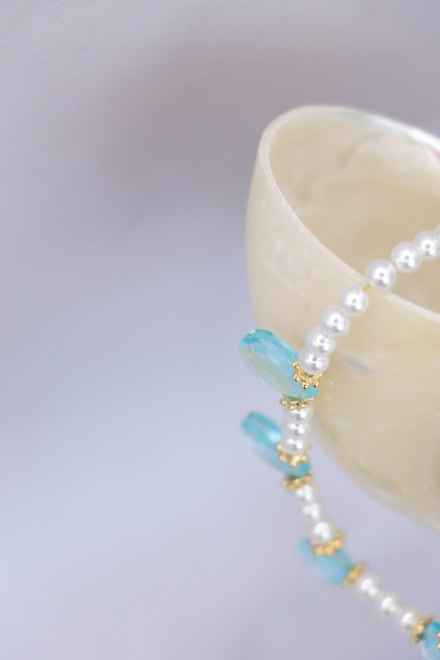 A close-up of a delicate beaded necklace draped over a cream-colored ceramic bowl. The necklace features white pearls, light blue faceted beads, and small gold spacer rings, set against a soft, out-of-focus lavender background.