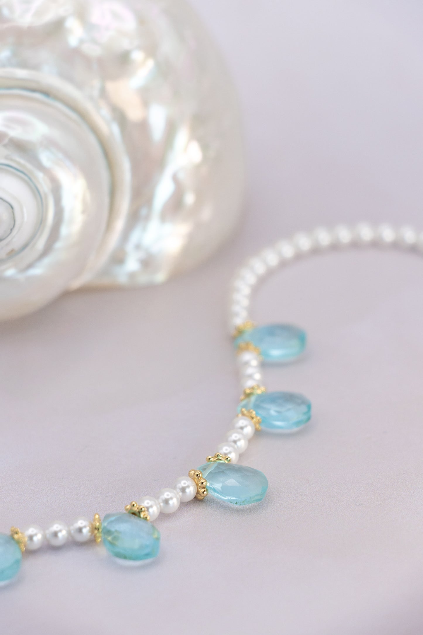A close-up of a beaded necklace with white pearls and light blue faceted beads, accented with small gold spacers, displayed on a pale pink surface next to a white seashell.