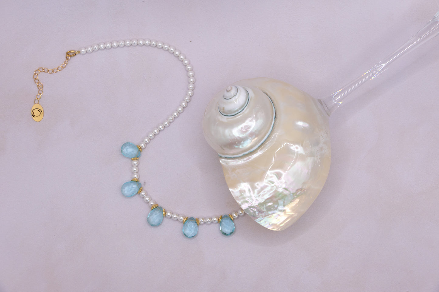 A pearl necklace with five light blue teardrop beads and gold-tone accents, displayed next to a large white seashell on a pale pink background.