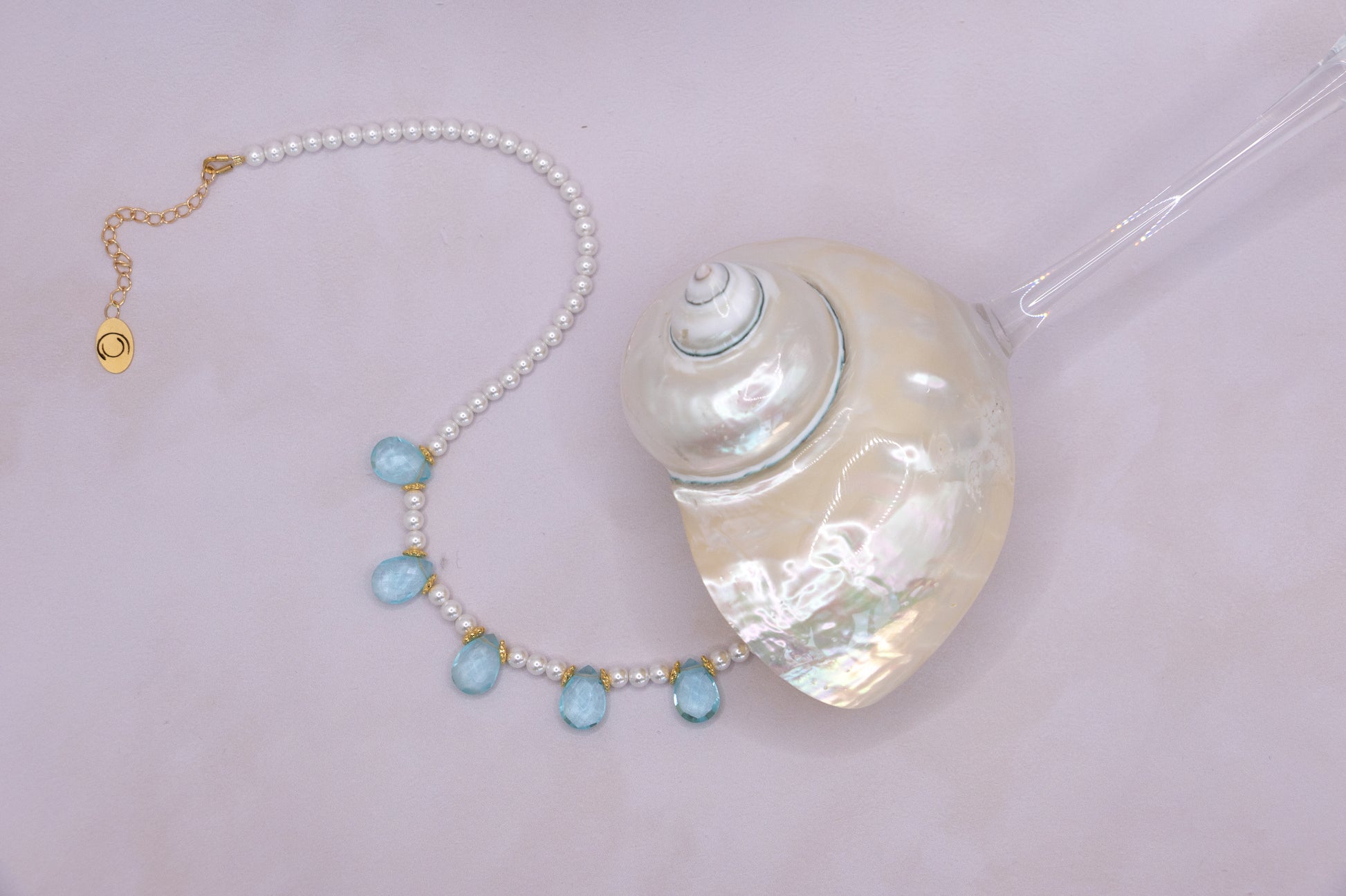 A pearl necklace with five light blue teardrop beads and gold-tone accents, displayed next to a large white seashell on a pale pink background.