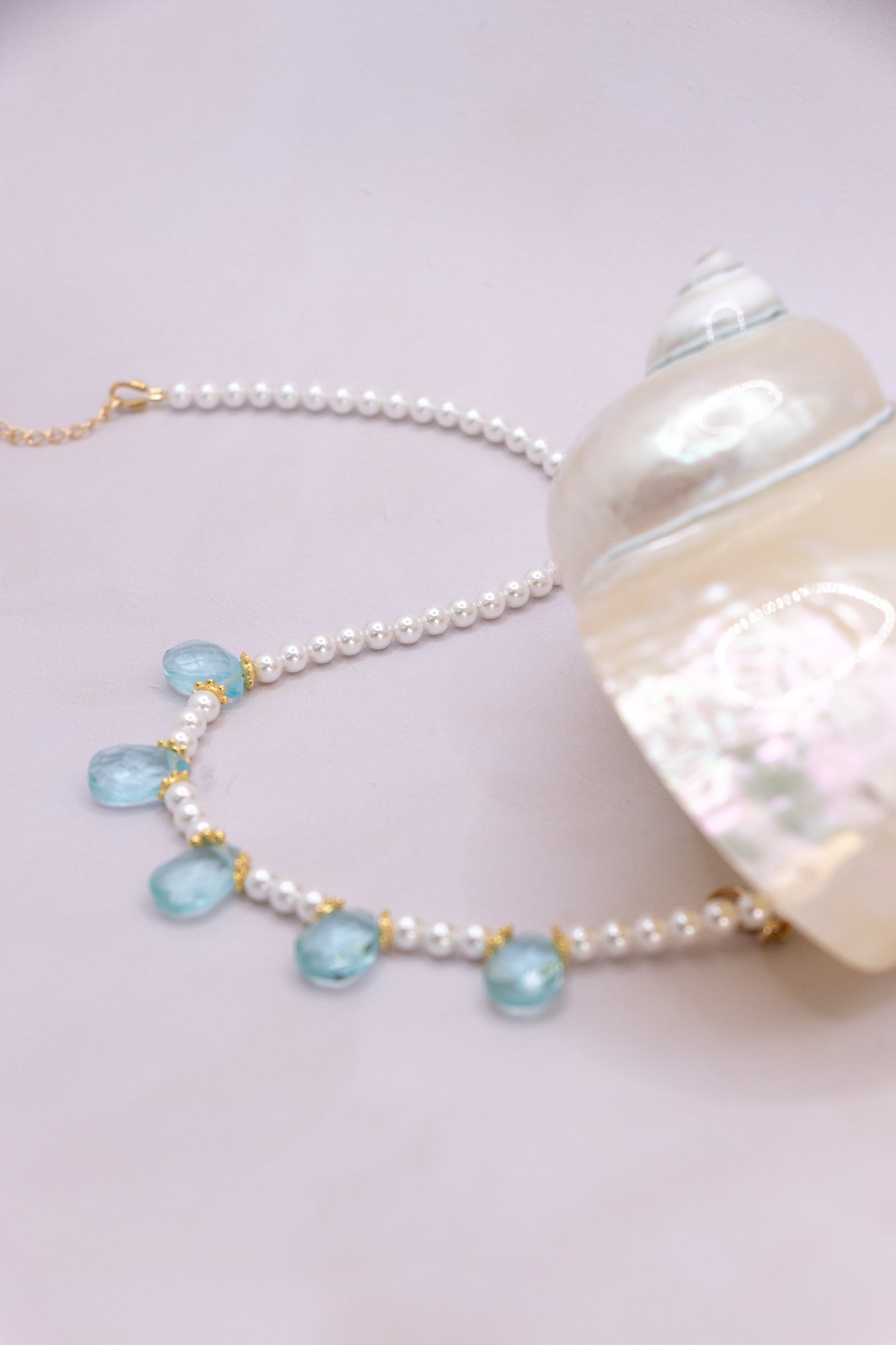 A pearl bead necklace with light blue teardrop-shaped gemstones and gold-tone accents, displayed next to a large white seashell on a pale pink background.