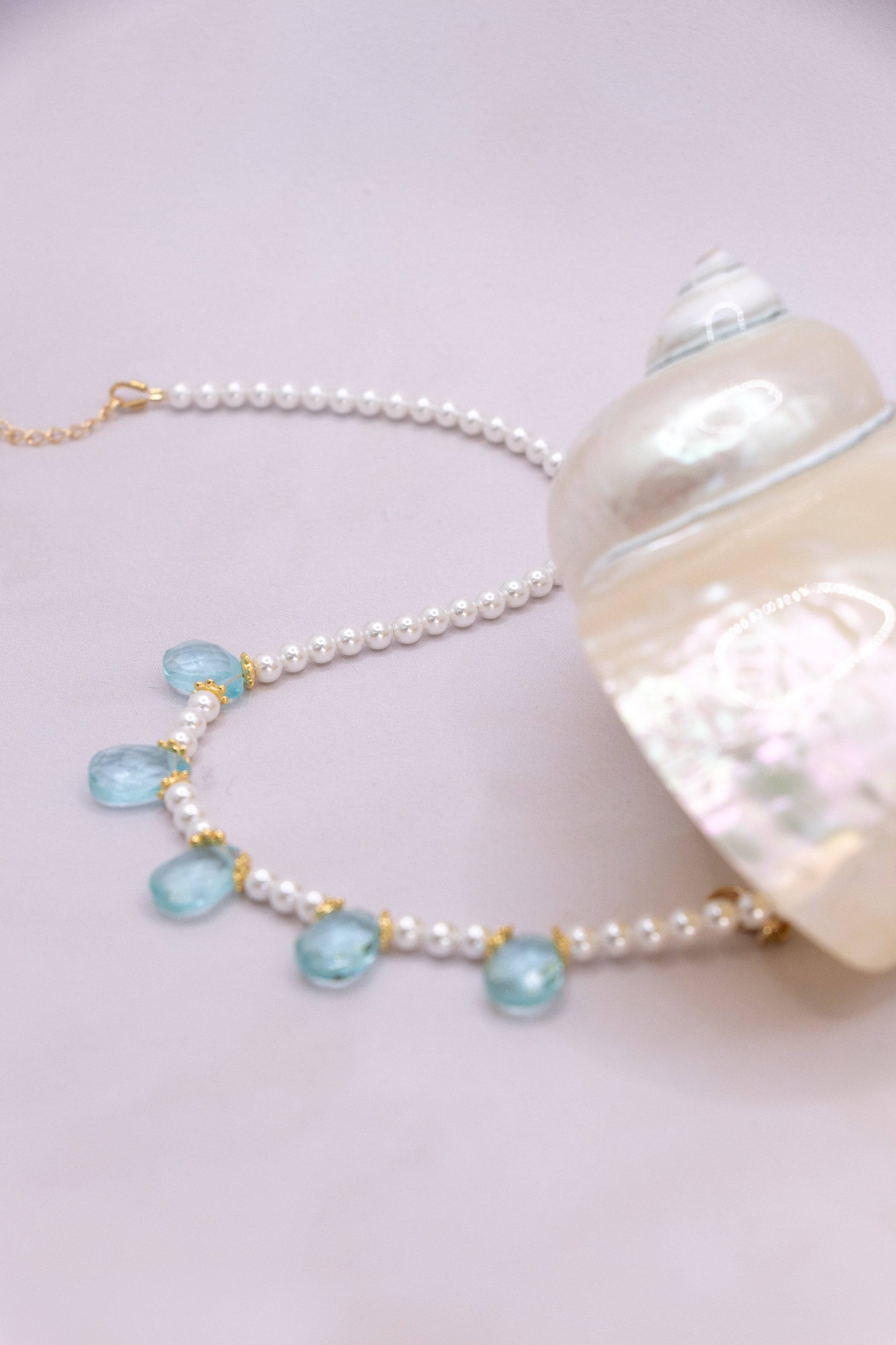 A pearl bead necklace with light blue teardrop-shaped gemstones and gold-tone accents, displayed next to a large white seashell on a pale pink background.