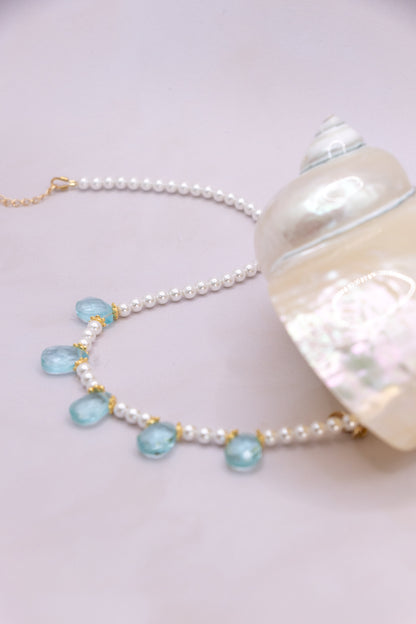A pearl bead necklace with light blue teardrop-shaped gemstones and gold-tone accents, displayed next to a large white seashell on a pale pink background.