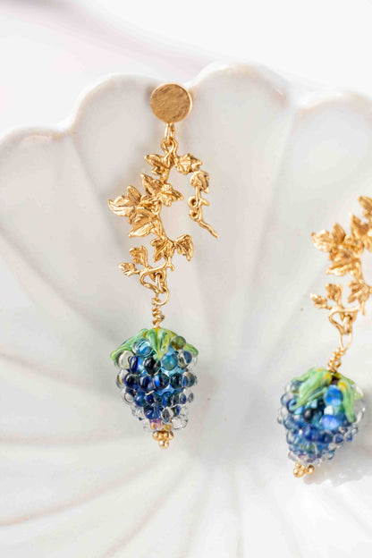 Gold and blue grape fruit earrings on a white shell background