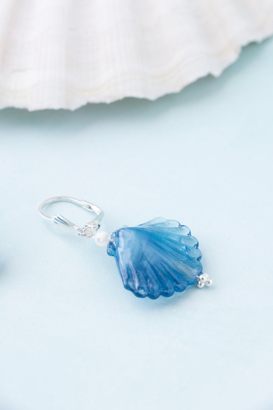 A silver-toned shell-shaped pendant charm with a translucent blue center, displayed on a light blue surface with a white seashell prop in the background.
