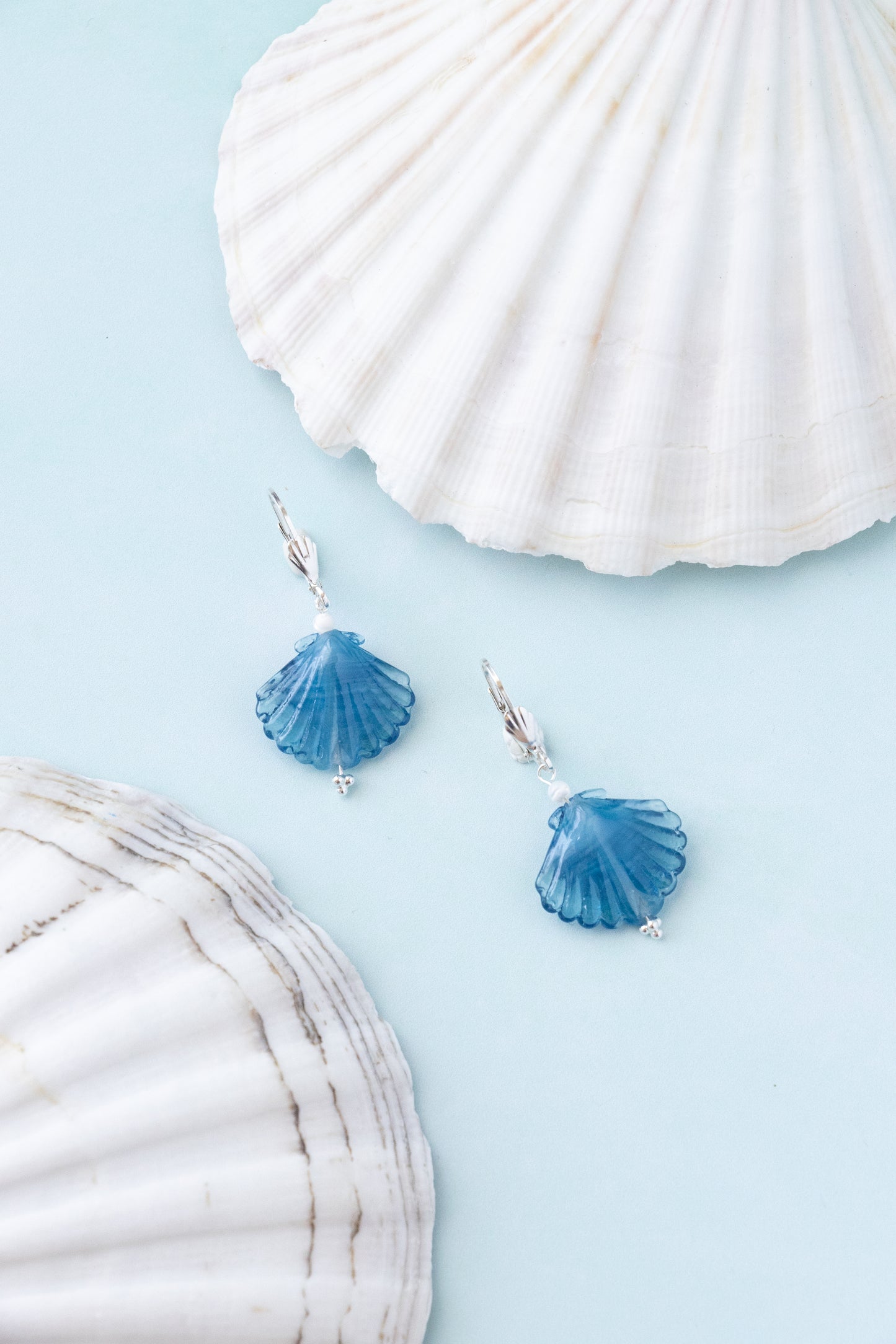 A pair of silver dangle earrings with blue scallop shell charms, styled on a light blue background with white seashells.