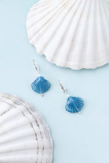 A pair of silver dangle earrings with blue scallop shell charms, styled on a light blue background with white seashells.