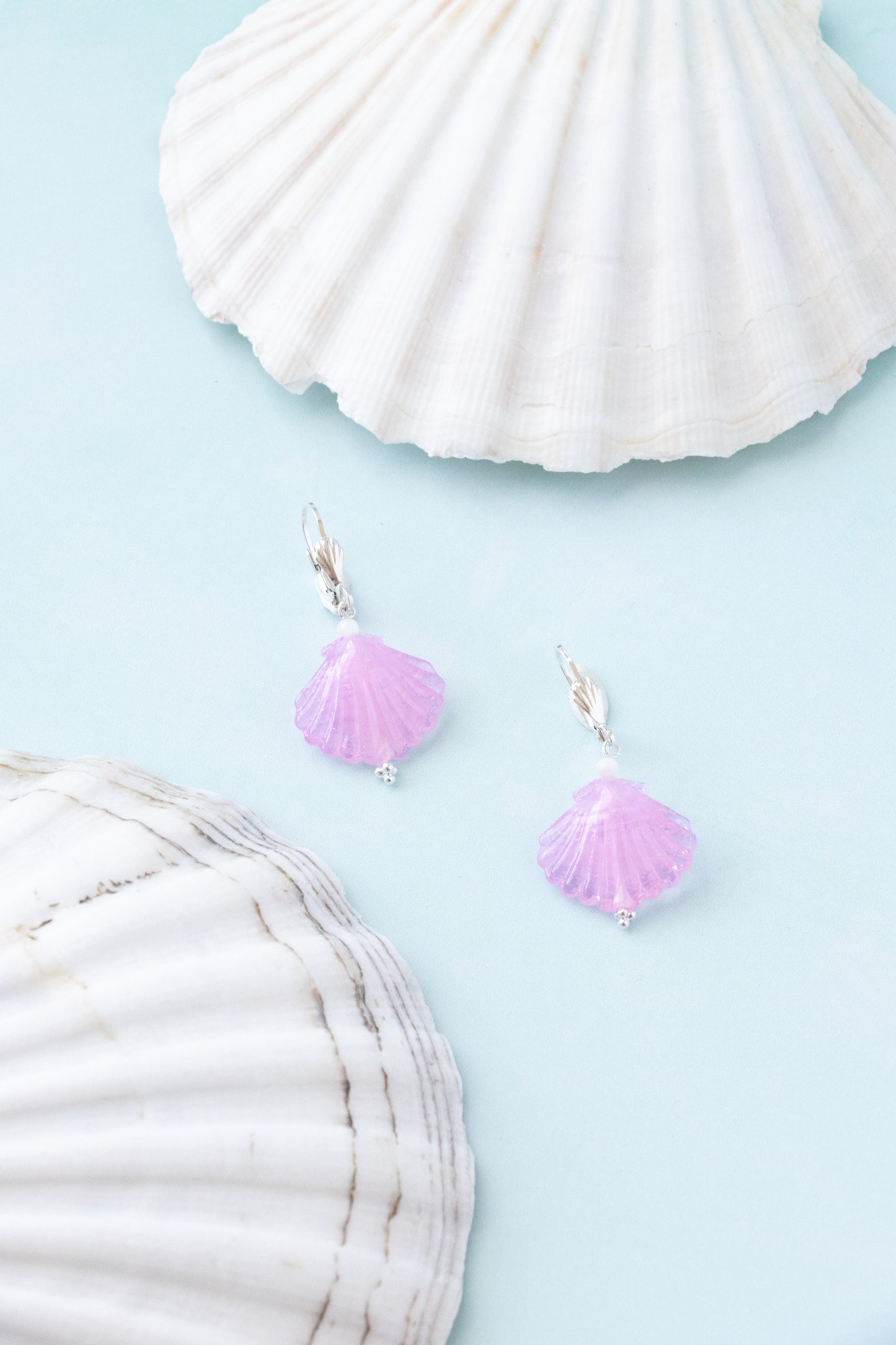 A pair of silver fishhook earrings with small lavender-pink seashell charms, displayed on a light blue background with decorative white seashells.