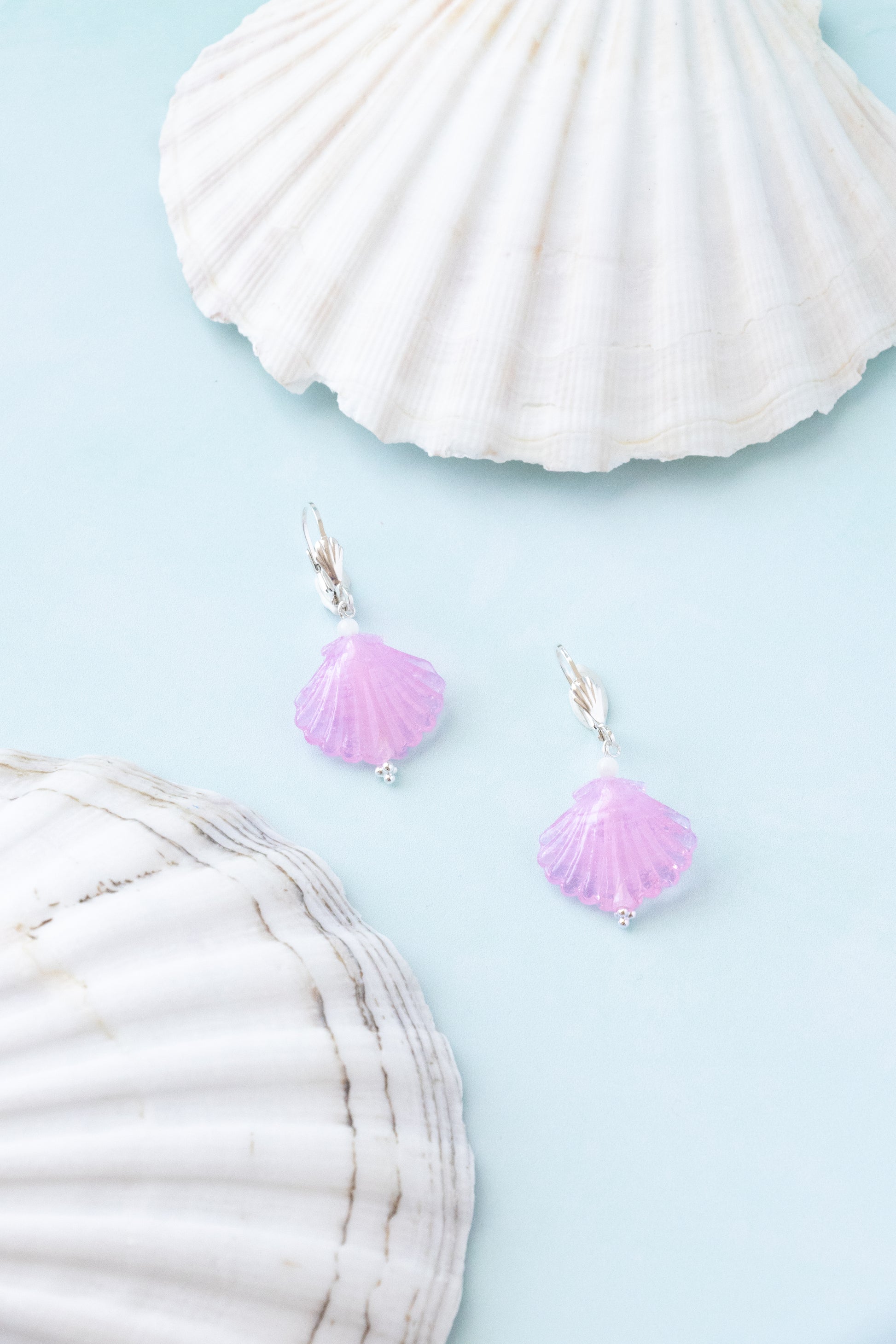A pair of silver fishhook earrings with small lavender-pink seashell charms, displayed on a light blue background with decorative white seashells.