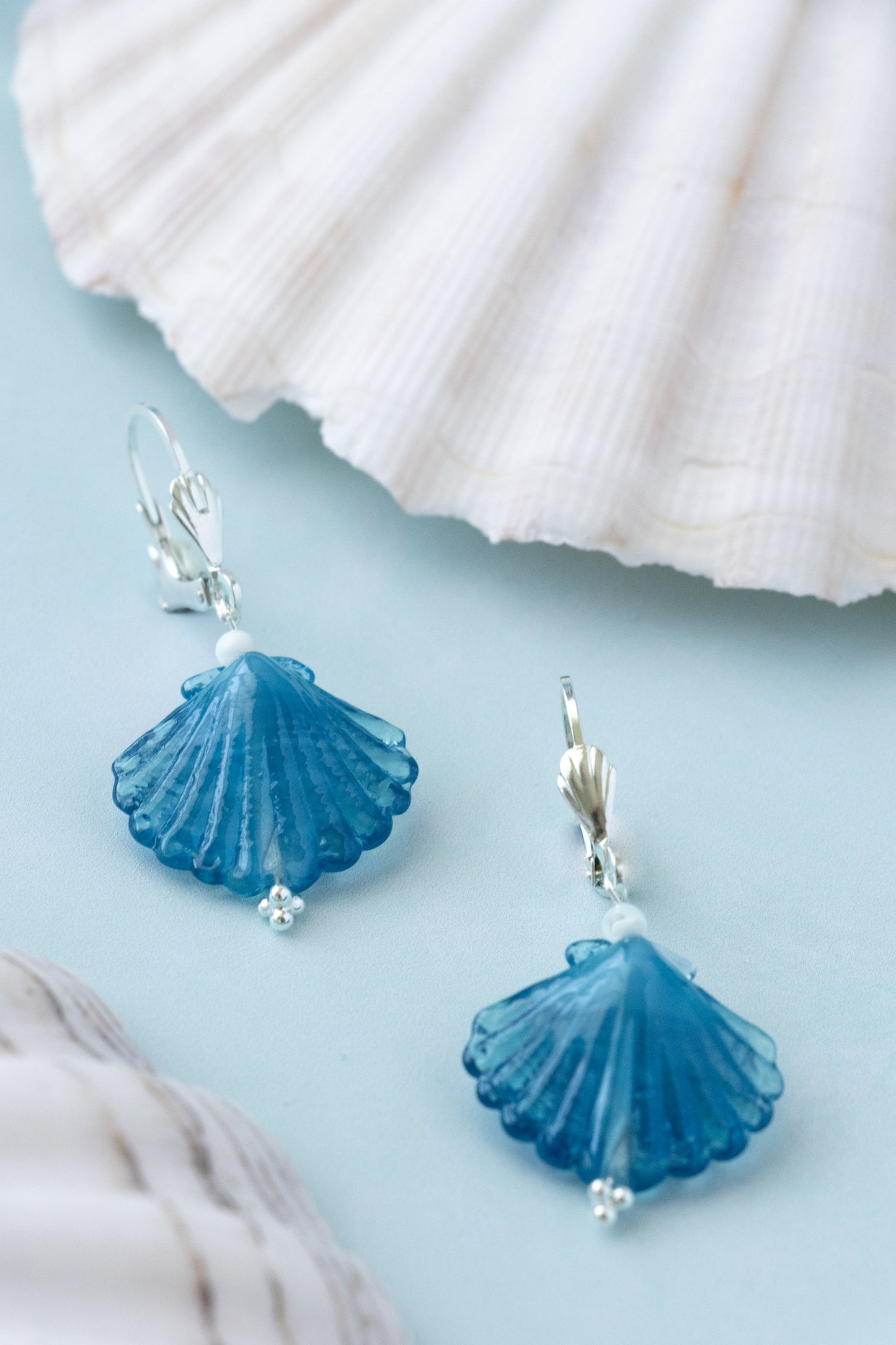 A pair of blue shell-shaped drop earrings with silver fishhook backs, displayed on a light blue surface with white seashells in the background.