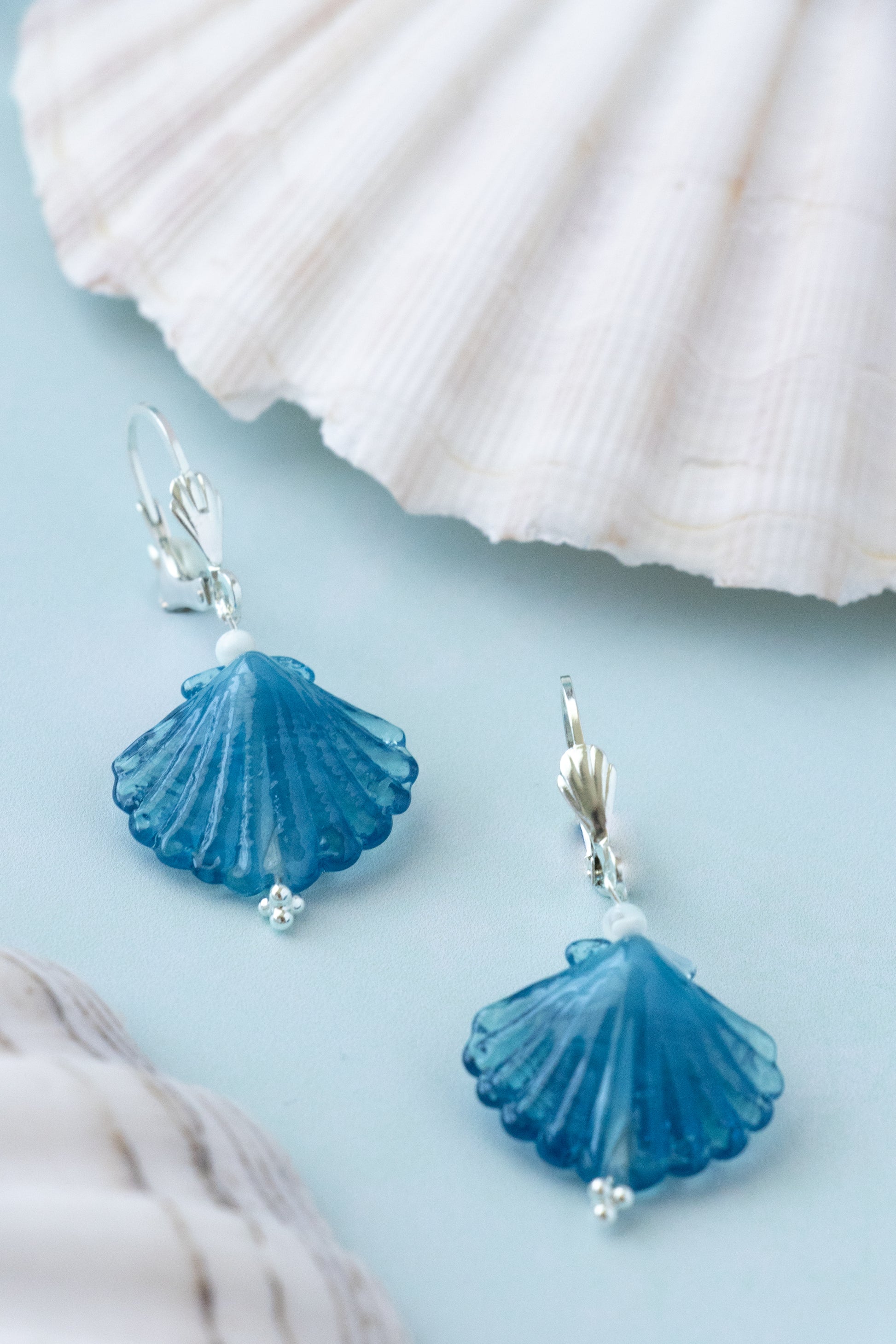 A pair of blue shell-shaped drop earrings with silver fishhook backs, displayed on a light blue surface with white seashells in the background.