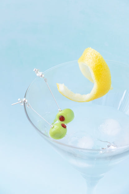 Silver dangling earrings shaped like green olives with red pimentos, hanging in a clear glass filled with ice and a lemon twist, against a light blue background.