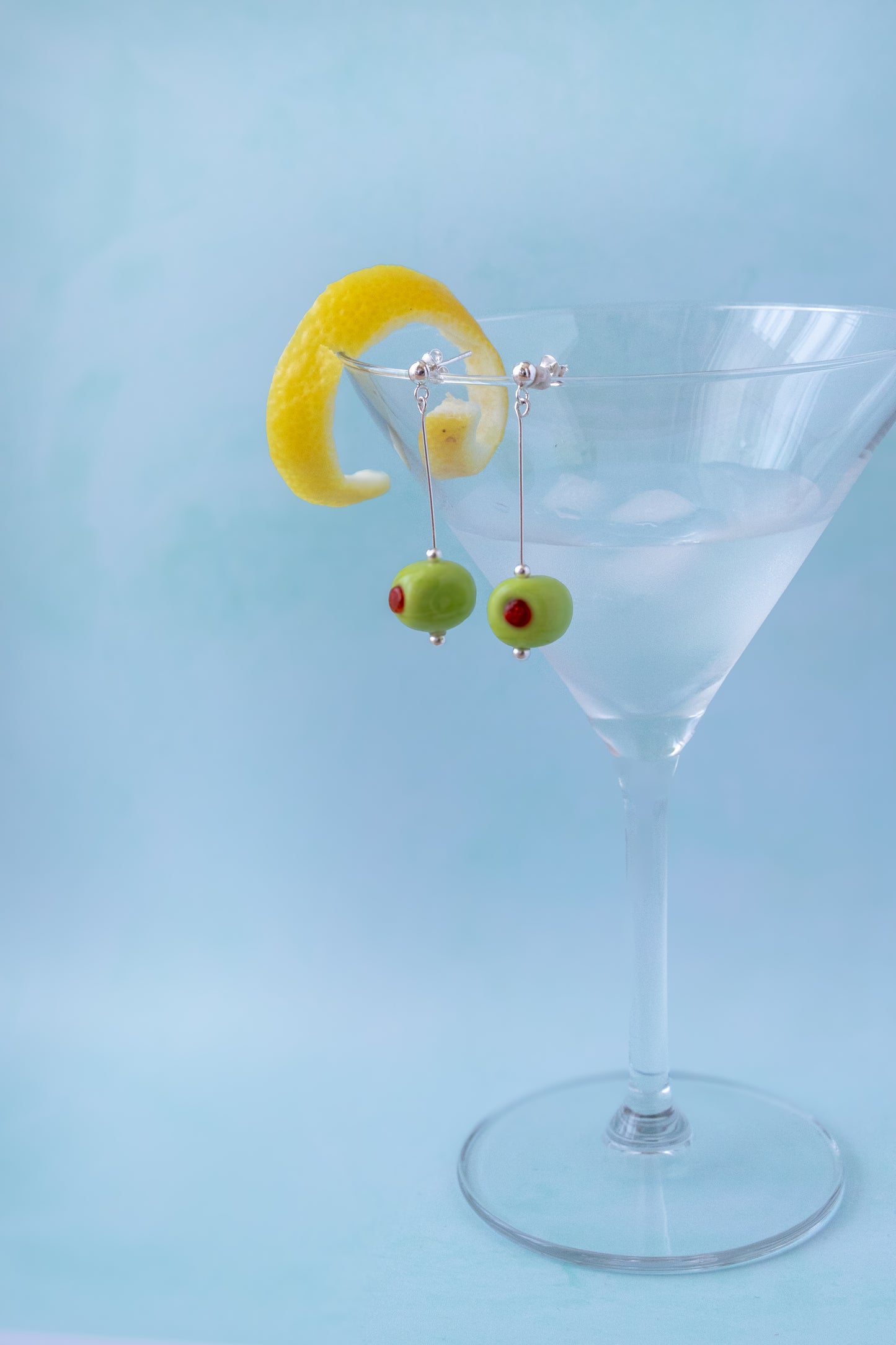 Silver dangling earrings shaped like green olives with red pimentos, hanging from a clear martini glass with a yellow lemon twist garnish against a light blue background.