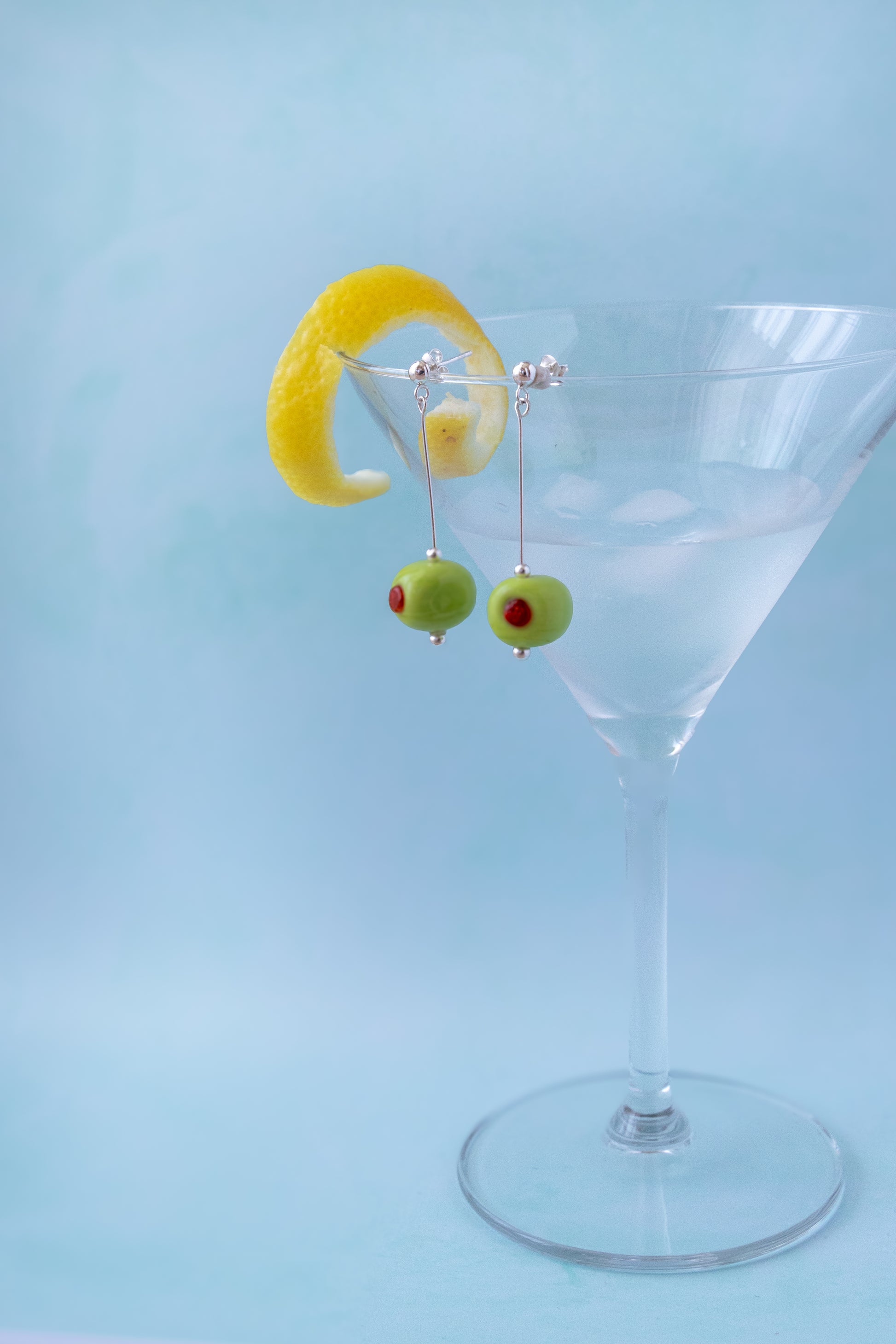 Silver dangling earrings shaped like green olives with red pimentos, hanging from a clear martini glass with a yellow lemon twist garnish against a light blue background.