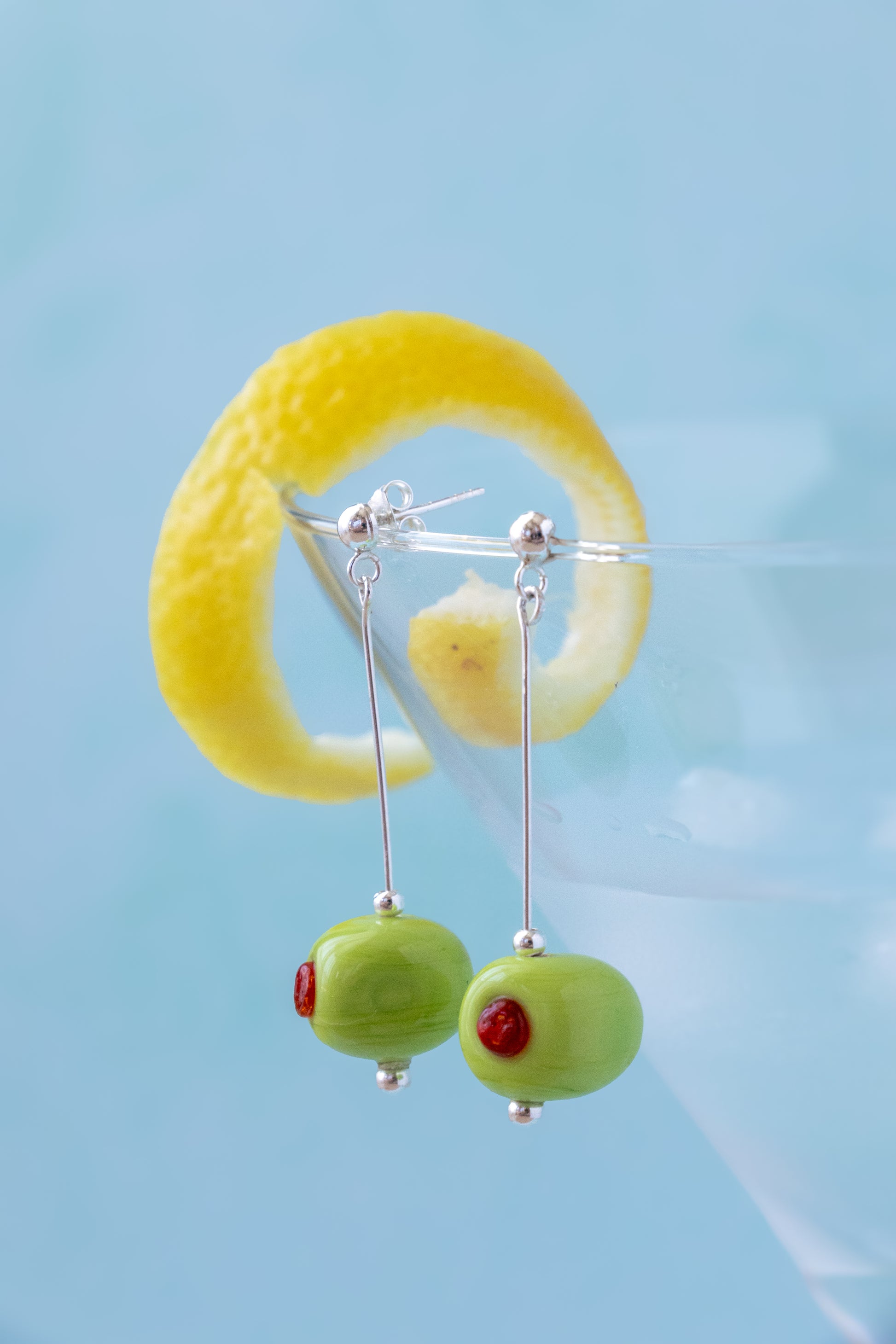 Green olive dangle earrings with small red eye details on silver hooks, displayed against a light blue background with a yellow lemon twist garnish.