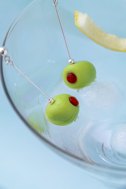 Green olive-shaped earrings with red centers hanging from silver wires, displayed inside a clear glass filled with ice and a lemon slice against a light blue background.