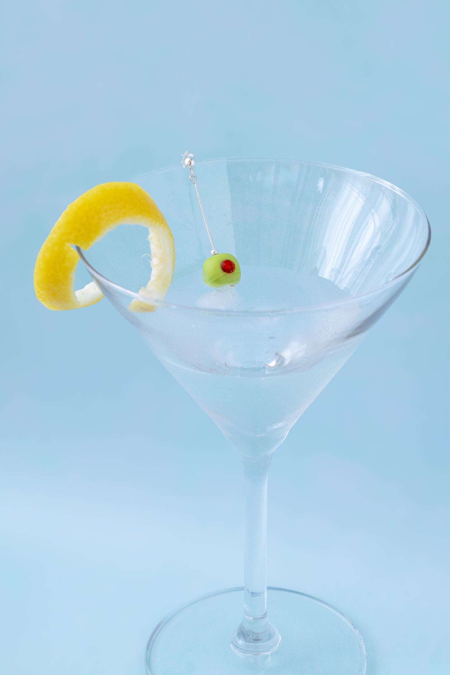 A clear martini glass on a light blue background, garnished with a curled yellow lemon peel and a small green olivette on a thin pick.