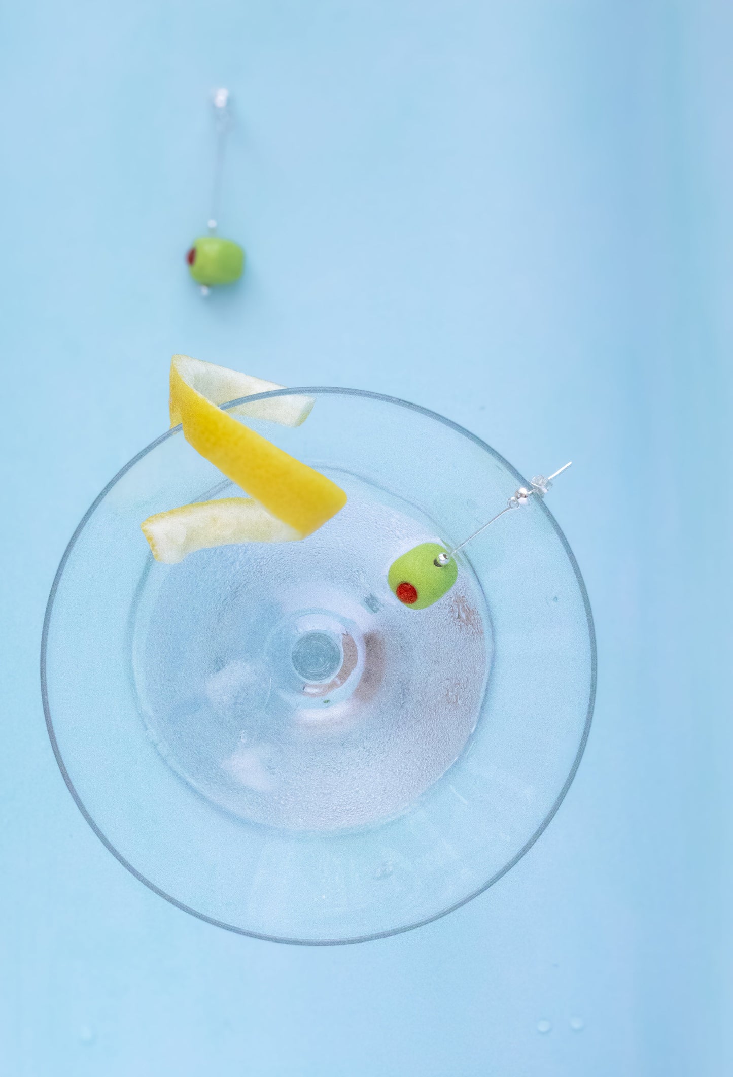 A top-down view of a clear glass coupe coupe-style drinkware on a light blue background, with a yellow lemon wedge and two green olives on silver skewers placed inside the glass.