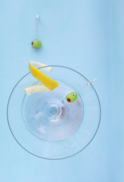 A top-down view of a clear glass coupe coupe-style drinkware on a light blue background, with a yellow lemon wedge and two green olives on silver skewers placed inside the glass.
