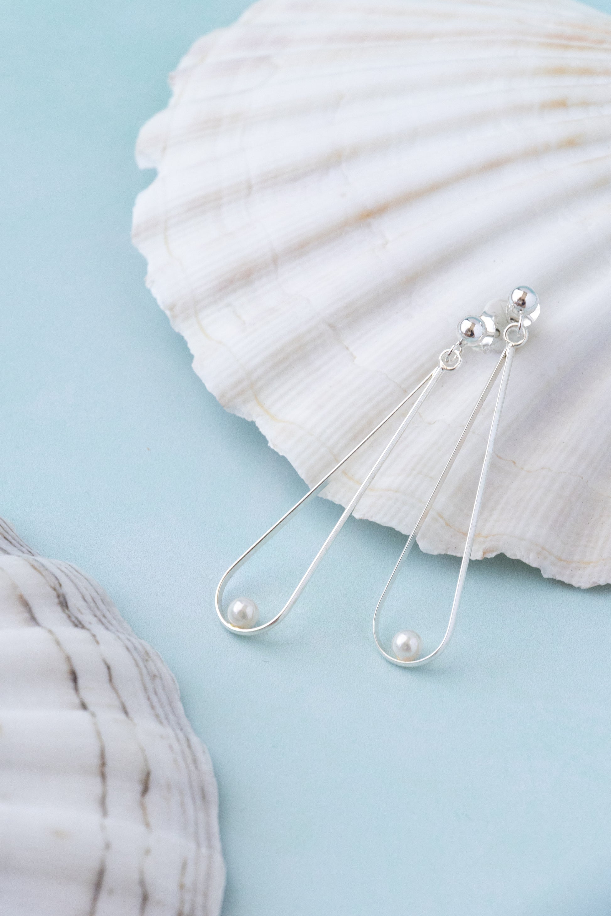 A pair of silver drop earrings with a small pearl on a blue summer background with a white seasshell