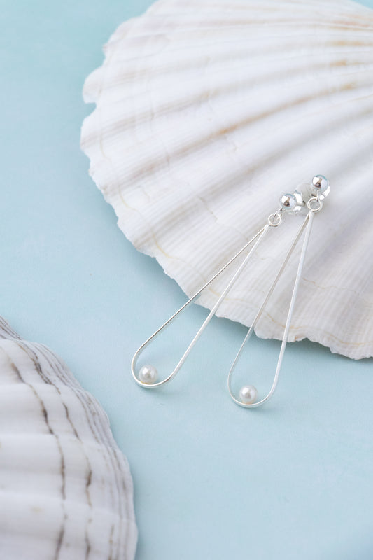 A pair of silver drop earrings with a small pearl on a blue summer background with a white seasshell