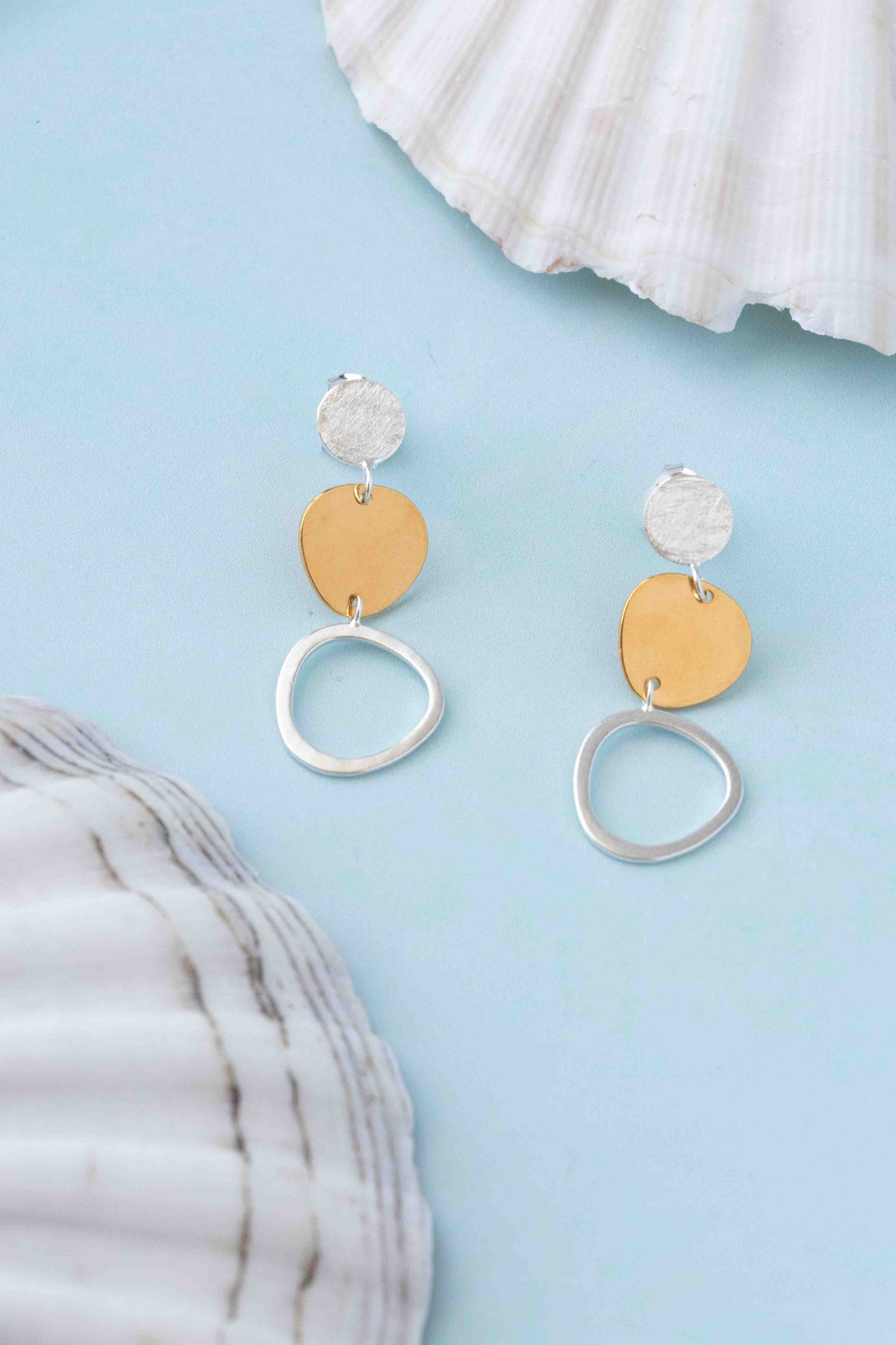 A pair of dangle earrings with textured silver studs, flat gold-tone circular discs, and dangling silver-tone open rings, displayed on a light blue background with white seashells as props.