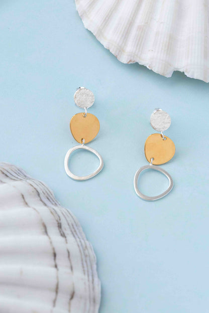 A pair of dangle earrings with textured silver studs, flat gold-tone circular discs, and dangling silver-tone open rings, displayed on a light blue background with white seashells as props.