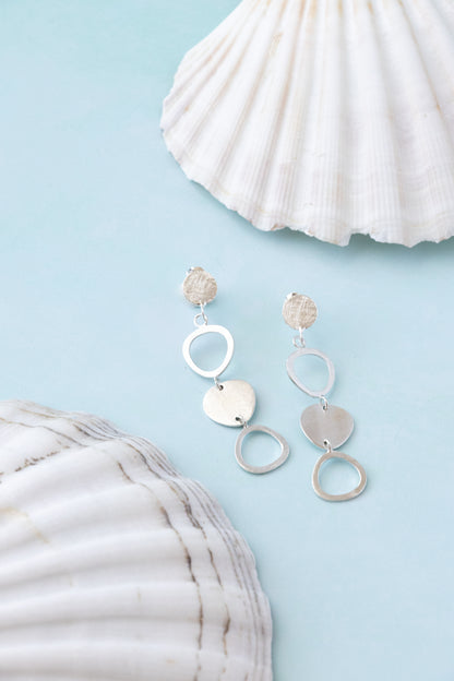 A pair of silver dangle earrings with textured circular and heart-shaped charms, displayed on a light blue background with white seashells as props.