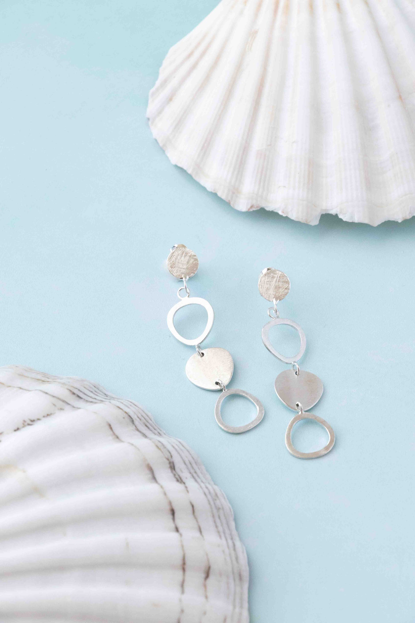 A pair of silver dangle earrings with circular and heart-shaped metal elements, displayed on a light blue background with white seashells as props.