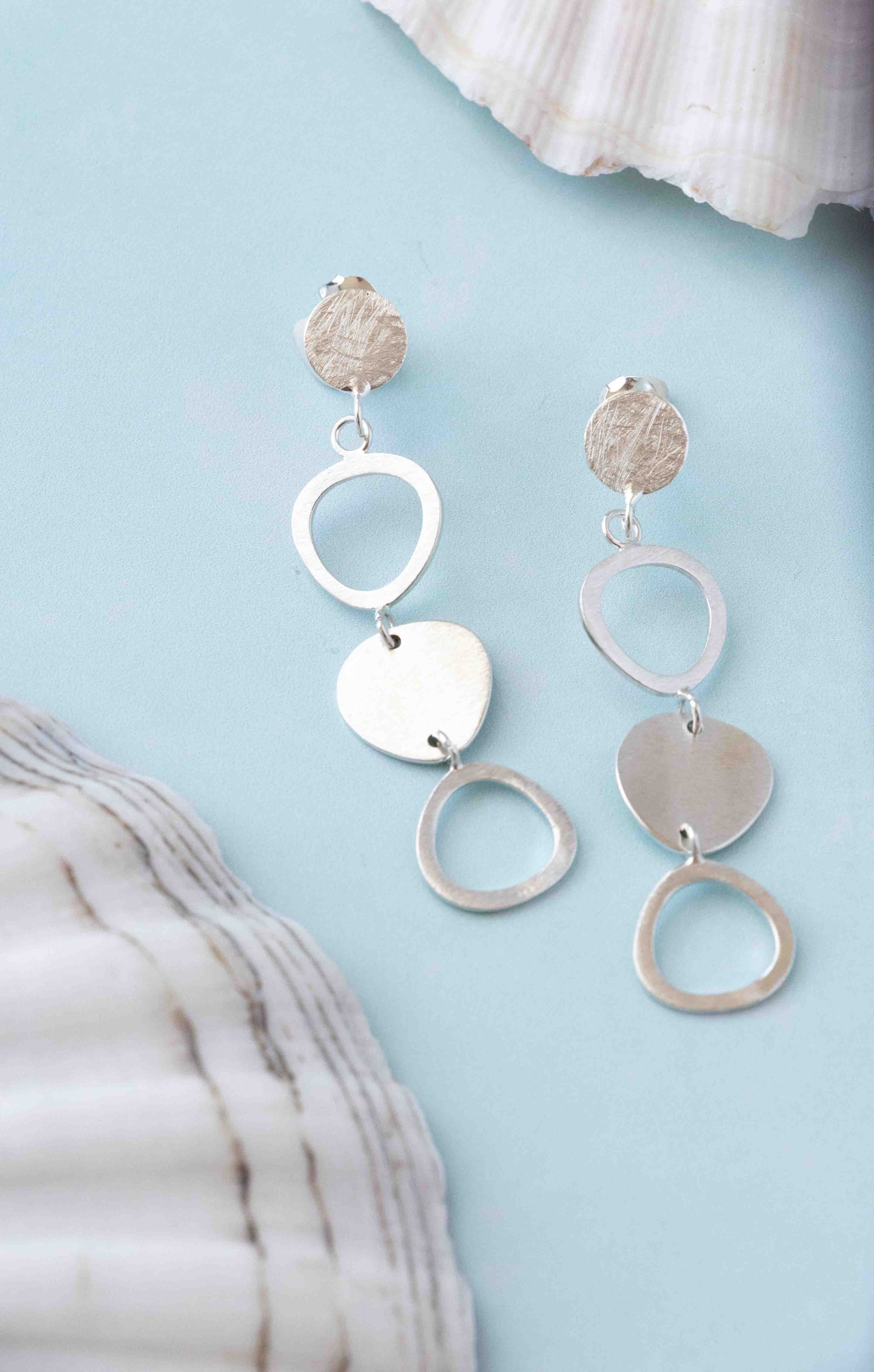 A pair of silver dangle earrings with circular, heart, and textured disc charms, displayed on a light blue background with seashell props.