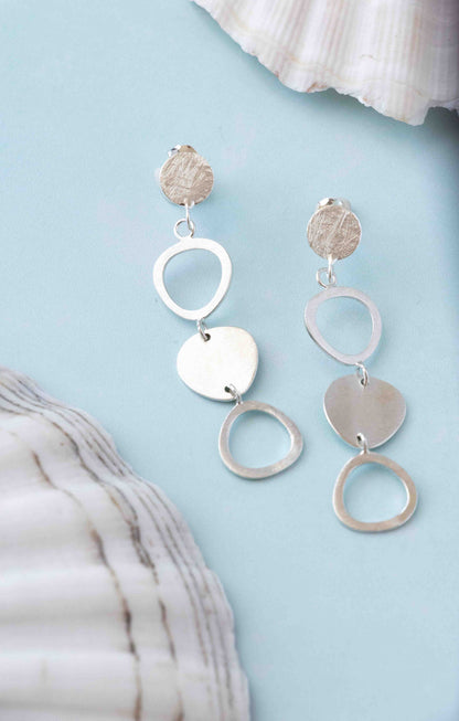 A pair of silver dangle earrings with circular, heart, and textured disc charms, displayed on a light blue background with seashell props.