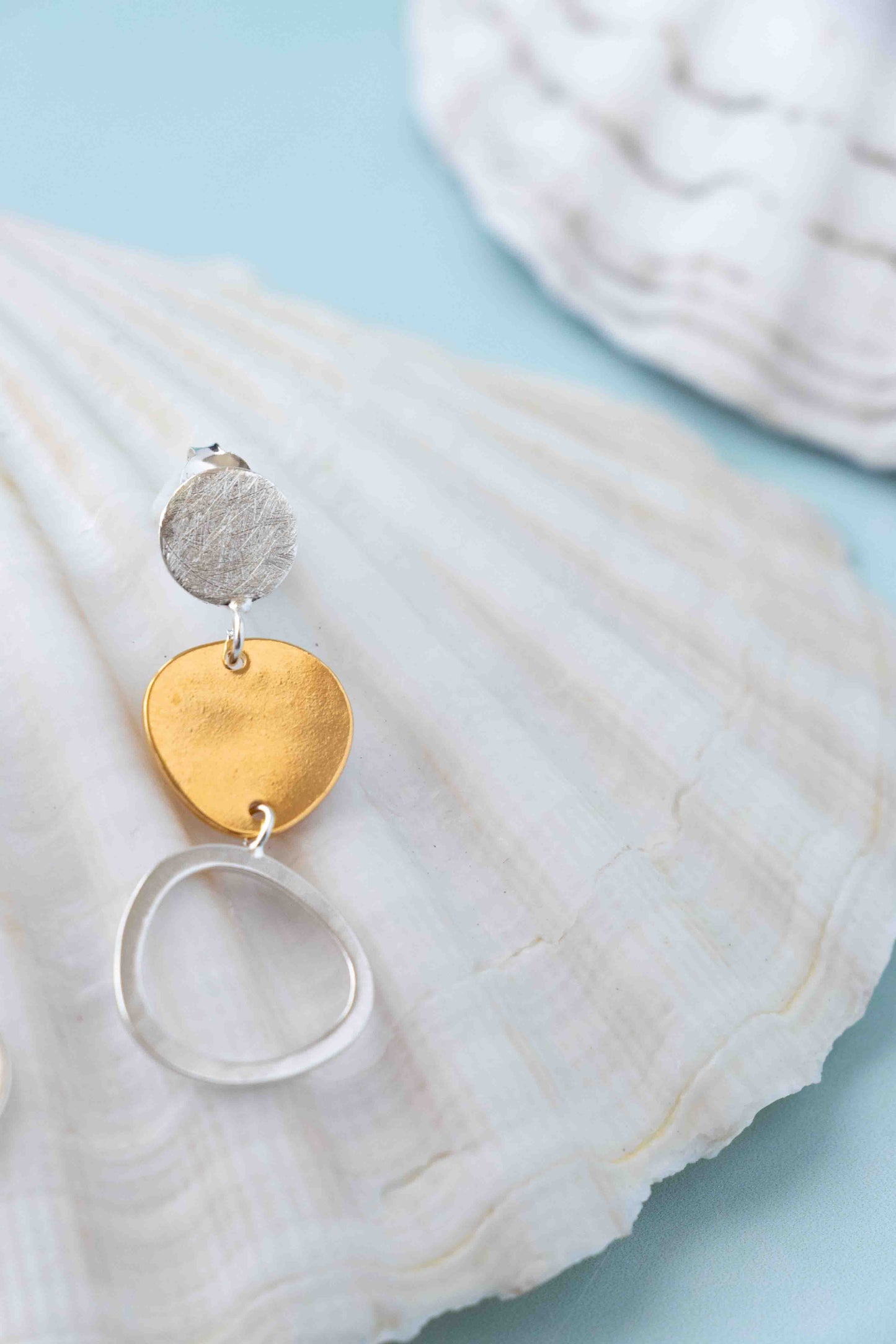 A pair of dangling earrings with textured silver and gold disc charms and a silver ring, displayed on a white seashell against a light blue background.