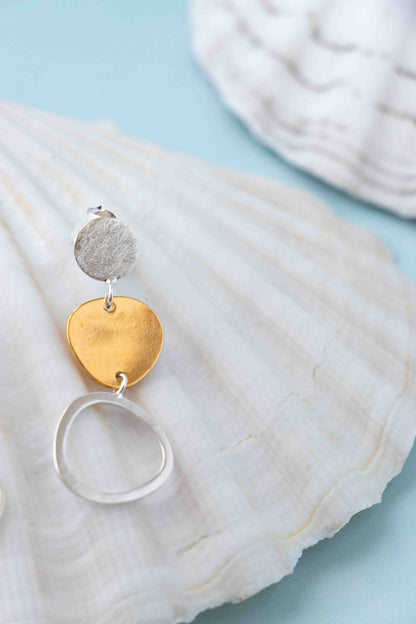 A pair of dangling earrings with textured silver and gold disc charms and a silver ring, displayed on a white seashell against a light blue background.