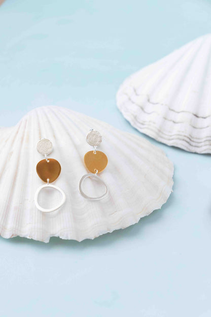 A pair of dangle earrings displayed on a white seashell against a light blue background, with a second shell blurred in the background. The earrings feature a textured silver top, a polished gold heart, and a silver ring.