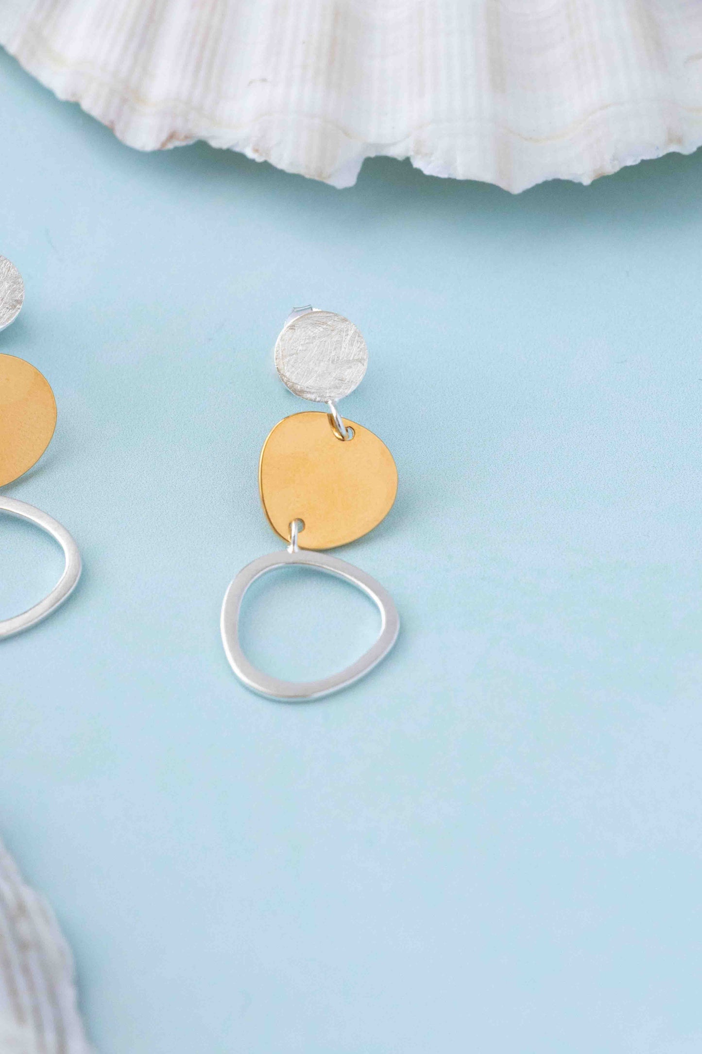 A pair of dangling earrings with a textured silver top disc, a flat gold heart-shaped disc, and a large open silver ring, displayed on a light blue surface with a white seashell prop.