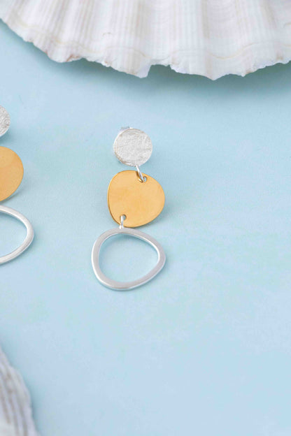 A pair of dangling earrings with a textured silver top disc, a flat gold heart-shaped disc, and a large open silver ring, displayed on a light blue surface with a white seashell prop.