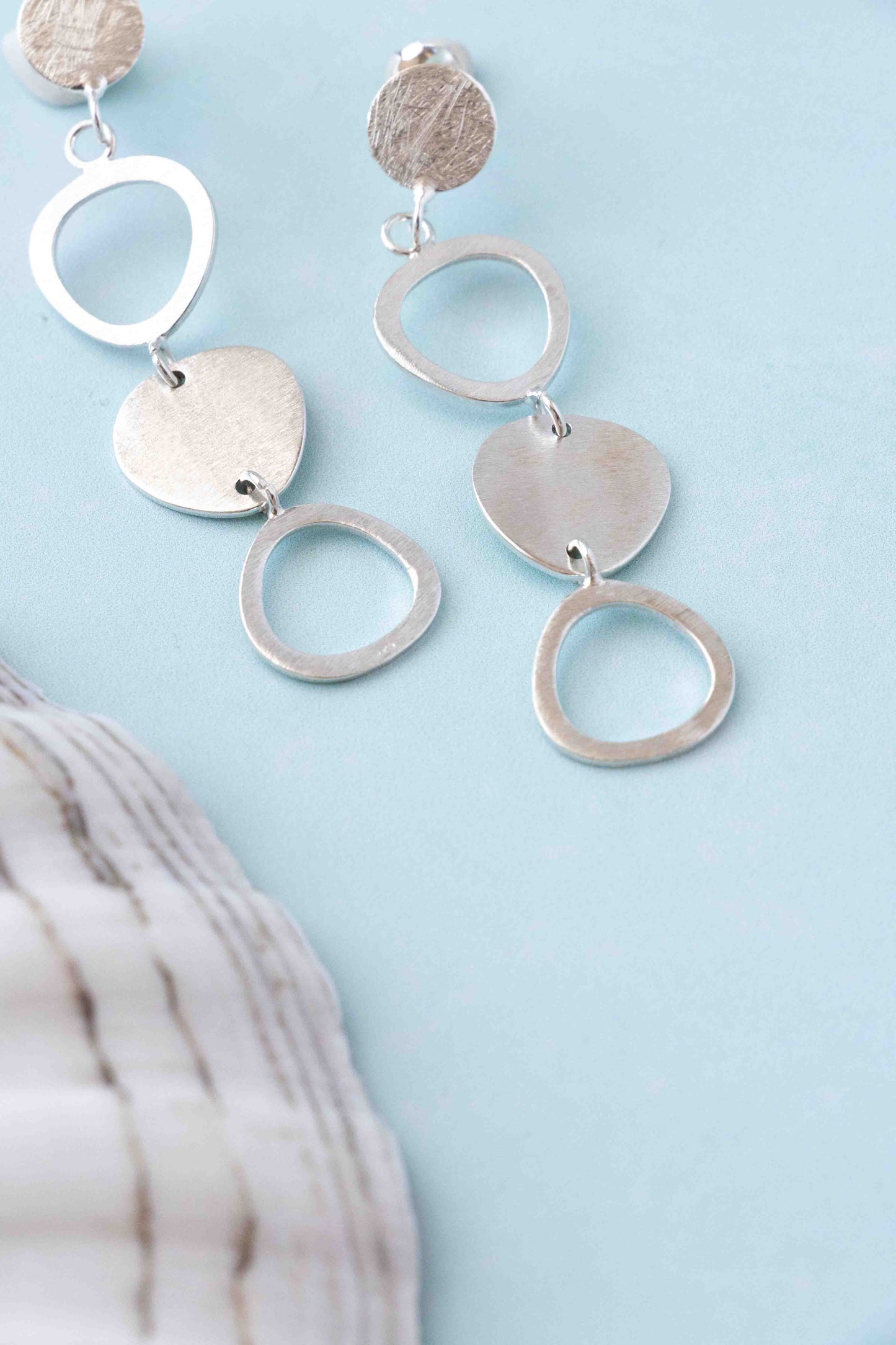 Sterling silver dangle earrings with textured circular and heart-shaped charms, displayed on a light blue background with a white seashell prop.