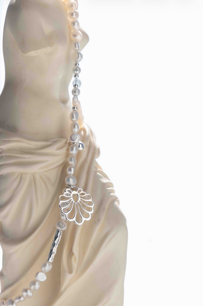 A close-up of a draped cream-colored statue wearing a long strand of white pearl beads with silver-toned accents, including a flower-shaped charm and a cylindrical bead.