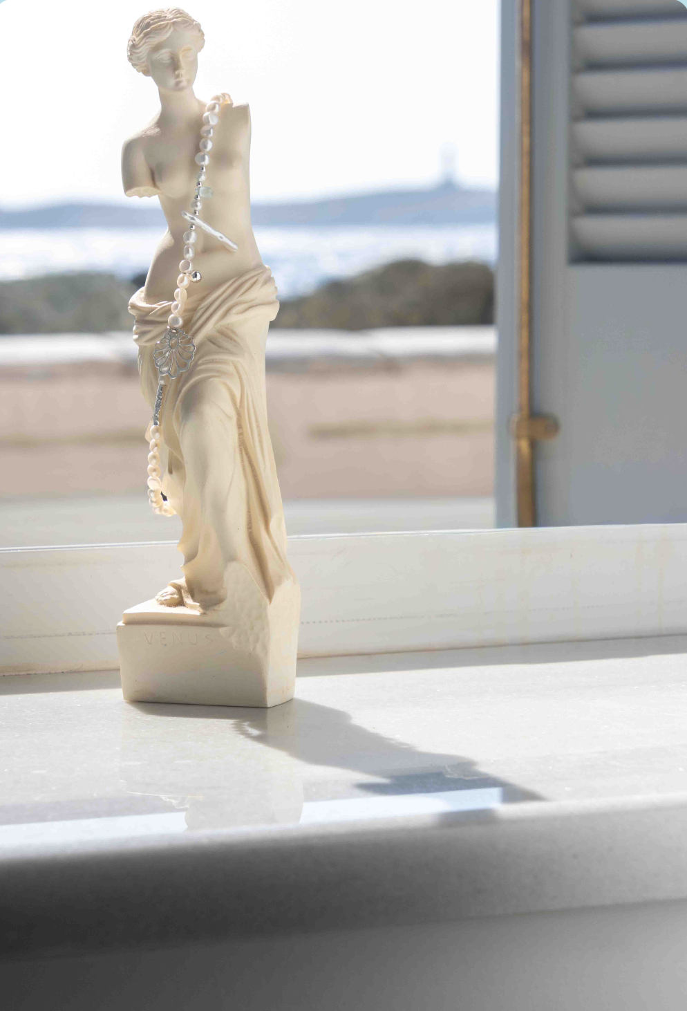 A small classical-style statue of a draped female figure in warm ivory/cream, placed on a light surface with a blurred window and outdoor view behind it.