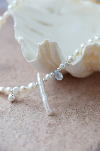 A close-up of a pearl and silver beaded necklace with a bar pendant and a small teardrop gemstone, draped over a white seashell on a sandy beige surface.