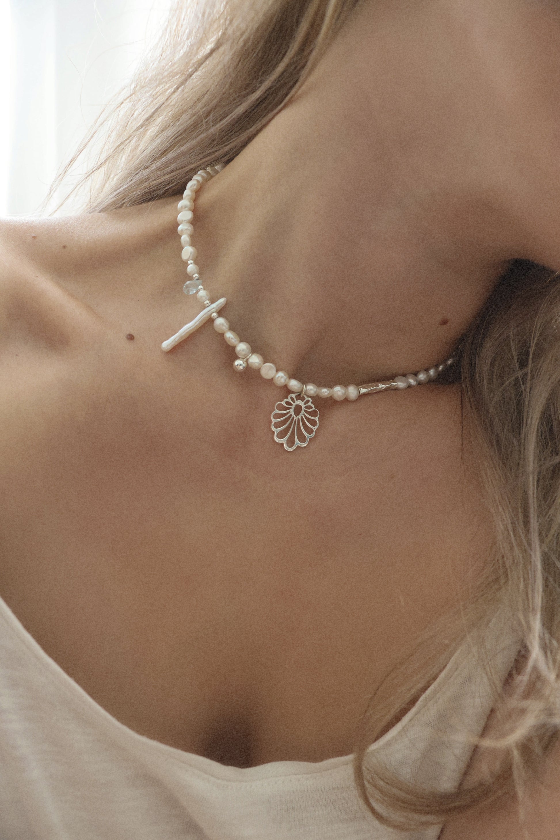A close-up lifestyle shot of a model wearing a delicate white pearl choker necklace with a silver cross and a silver filigree leaf pendant.